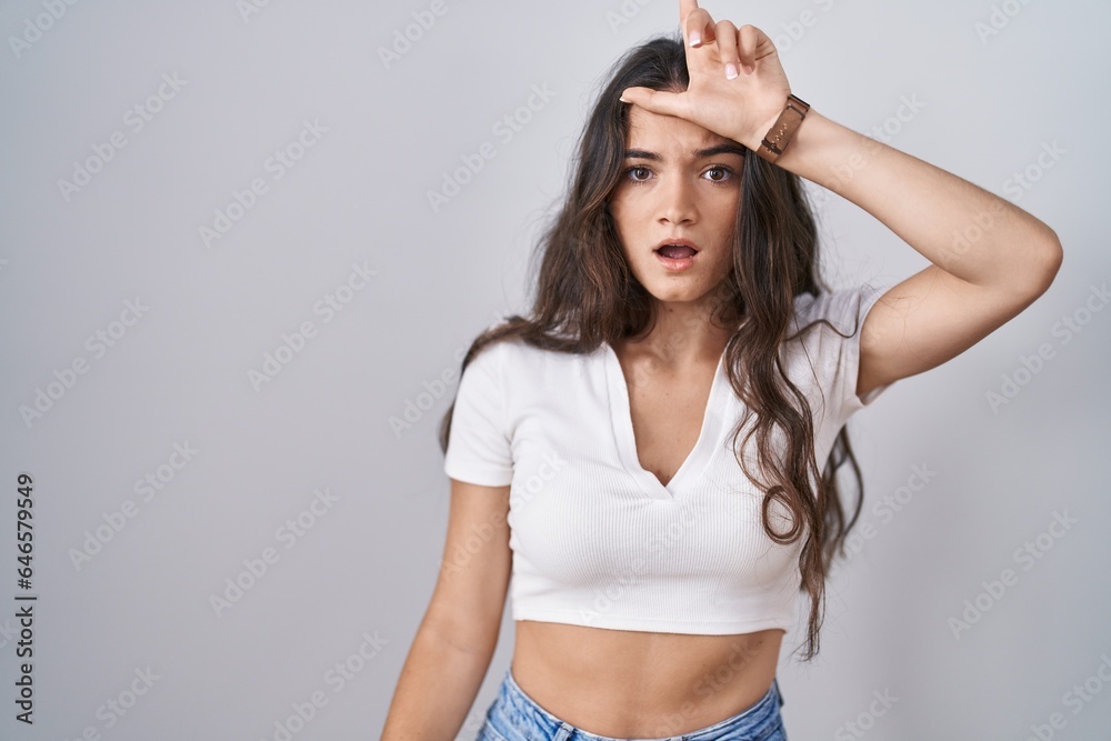 Obraz premium Young teenager girl standing over white background making fun of people with fingers on forehead doing loser gesture mocking and insulting.