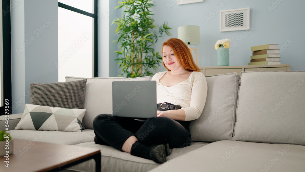 Young redhead woman using laptop sitting on sofa at home Young redhead woman using laptop sitting on sofa at home