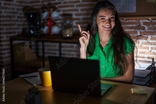 Young teenager girl working at the office at night with a big smile on face, pointing with hand and finger to the side looking at the camera.