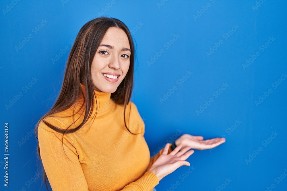 Fototapeta premium Young brunette woman standing over blue background inviting to enter smiling natural with open hand