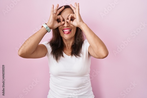 Middle age brunette woman standing over pink background doing ok gesture like binoculars sticking tongue out, eyes looking through fingers. crazy expression.