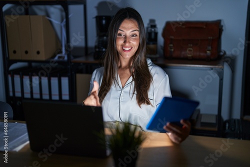 Young brunette woman working at the office at night smiling friendly offering handshake as greeting and welcoming. successful business. photo