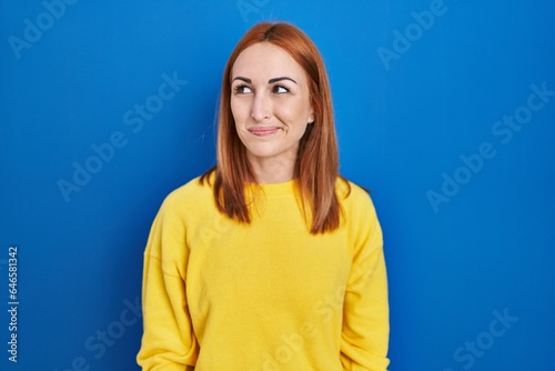 Young woman standing over blue background smiling looking to the side and staring away thinking.
