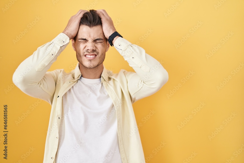 Young hispanic man standing over yellow background suffering from headache desperate and stressed because pain and migraine. hands on head. Young hispanic man standing over yellow background suffering from headache desperate and stressed because pain and migraine. hands on head.