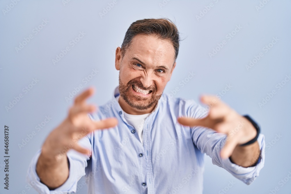Middle age caucasian man standing over blue background shouting frustrated with rage, hands trying to strangle, yelling mad Middle age caucasian man standing over blue background shouting frustrated with rage, hands trying to strangle, yelling mad