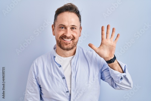 Middle age caucasian man standing over blue background showing and pointing up with fingers number five while smiling confident and happy. © Krakenimages.com
