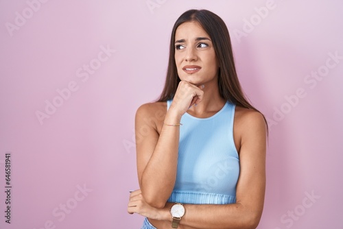 Young brunette woman standing over pink background thinking worried about a question, concerned and nervous with hand on chin