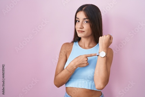 Young brunette woman standing over pink background in hurry pointing to watch time, impatience, looking at the camera with relaxed expression © Krakenimages.com