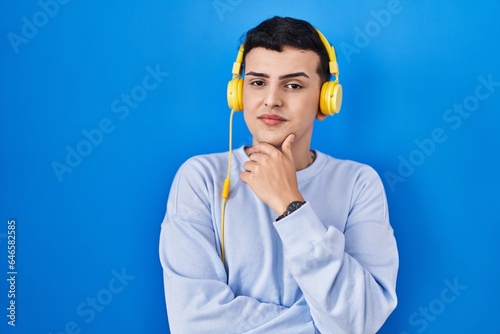 Non binary person listening to music using headphones looking confident at the camera smiling with crossed arms and hand raised on chin. thinking positive.