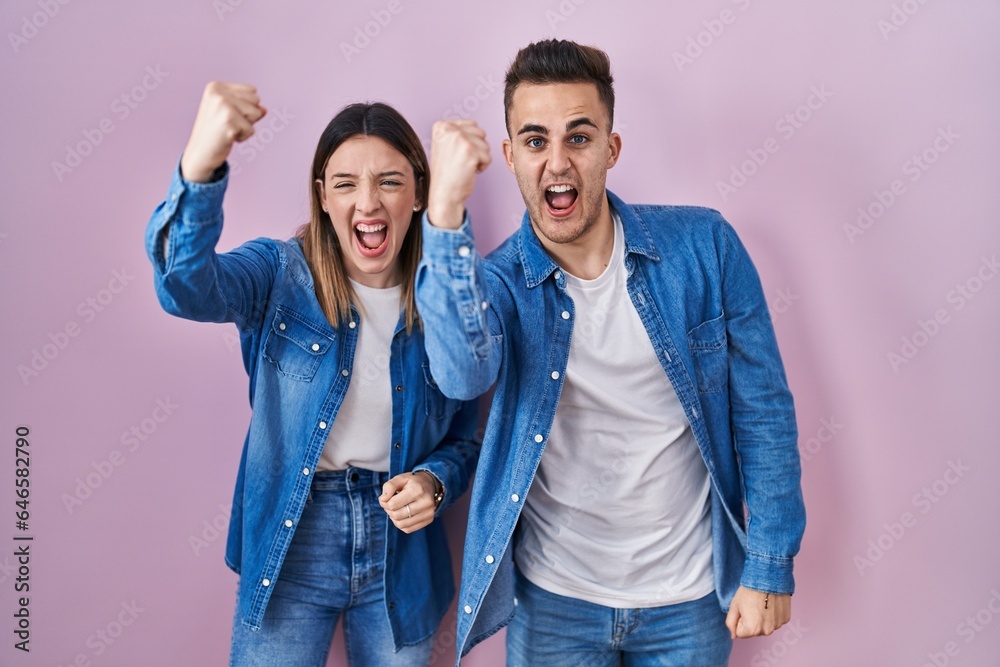 Young hispanic couple standing over pink background angry and mad raising fist frustrated and furious while shouting with anger. rage and aggressive concept. Young hispanic couple standing over pink background angry and mad raising fist frustrated and furious while shouting with anger. rage and aggressive concept.