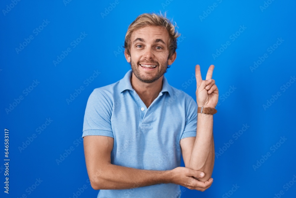 Naklejka premium Caucasian man standing over blue background smiling with happy face winking at the camera doing victory sign with fingers. number two.