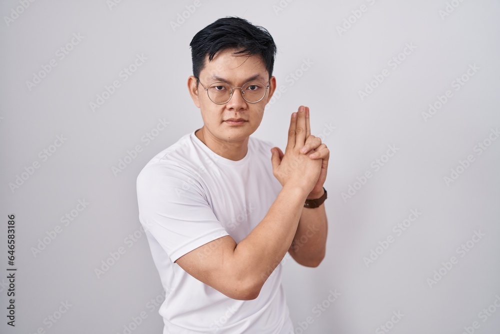 Fototapeta premium Young asian man standing over white background holding symbolic gun with hand gesture, playing killing shooting weapons, angry face
