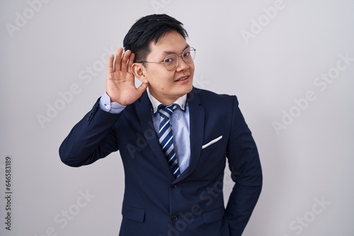 Young asian man wearing business suit and tie smiling with hand over ear listening an hearing to rumor or gossip. deafness concept.