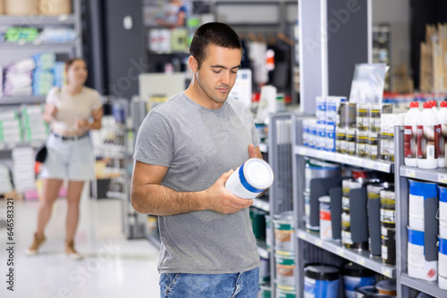 In store, male buyer doubts choice and examines product, paint jar studies detailed information on packaging, label. Repair and construction items, paint and varnish products, accessories, supermarke.