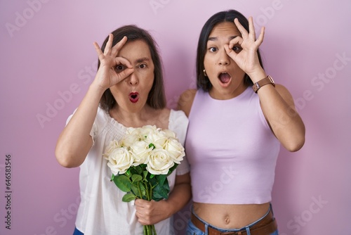 Hispanic mother and daughter holding bouquet of white flowers doing ok gesture shocked with surprised face, eye looking through fingers. unbelieving expression.