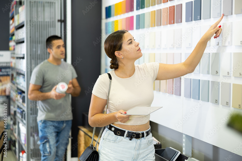 Young woman stand near showcase with samples of colors, textures of paint and varnish materials and choose best cover option. Repair and construction items, accessories, supermarket