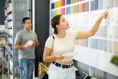 Young woman stand near showcase with samples of colors, textures of paint and varnish materials and choose best cover option. Repair and construction items, accessories, supermarket
