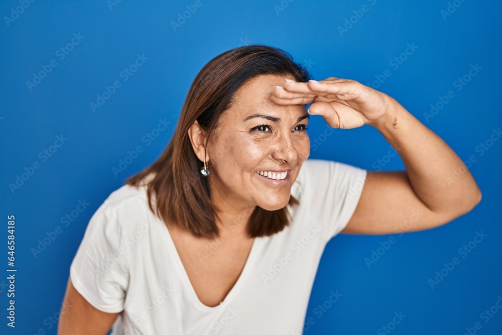 Hispanic mature woman standing over blue background very happy and smiling looking far away with hand over head. searching concept. Hispanic mature woman standing over blue background very happy and smiling looking far away with hand over head. searching concept.