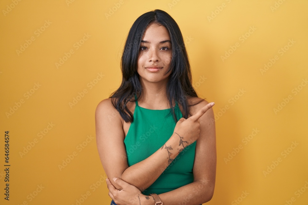 Brunette woman standing over yellow background pointing with hand finger to the side showing advertisement, serious and calm face Brunette woman standing over yellow background pointing with hand finger to the side showing advertisement, serious and calm face