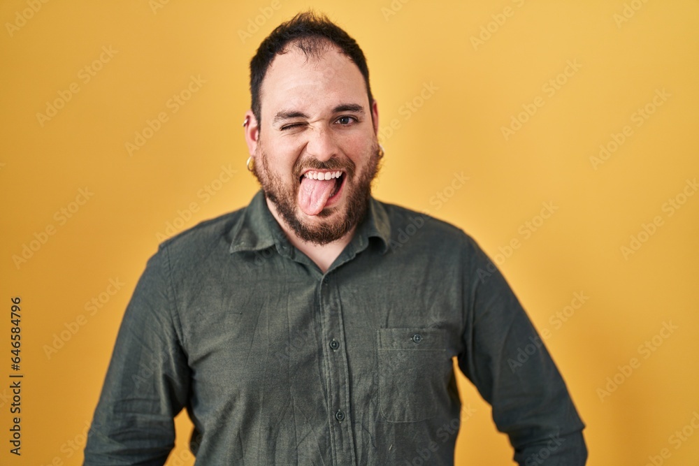 Plus size hispanic man with beard standing over yellow background sticking tongue out happy with funny expression. emotion concept. Plus size hispanic man with beard standing over yellow background sticking tongue out happy with funny expression. emotion concept.