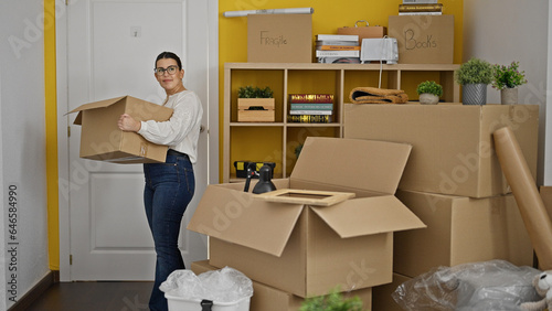 Young beautiful hispanic woman walking in holding package at new home