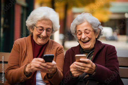 Two senior women sitting outdoors looking at their smart phones and laughing.