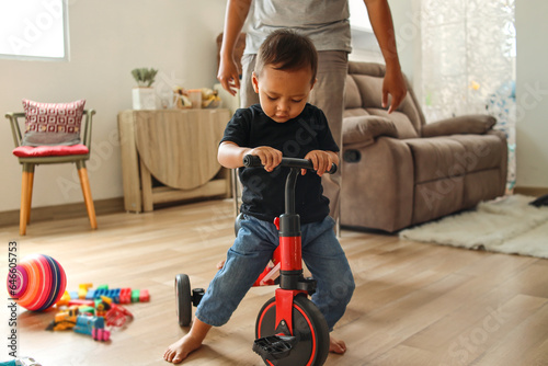 Adorable Asian little boy playing tricyle at home while his father watching and standing to protect him photo