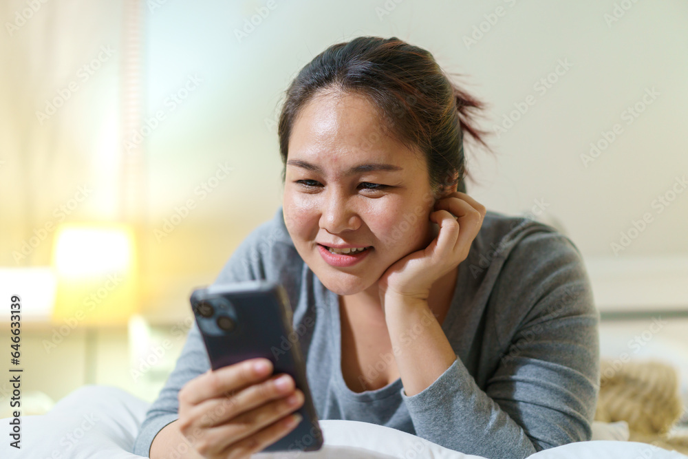 Asian oversize fat woman lying on the bed at night and chatting or messaging with a friends on smartphone before getting sleep. Asian oversize fat woman lying on the bed at night and chatting or messaging with a friends on smartphone before getting sleep.