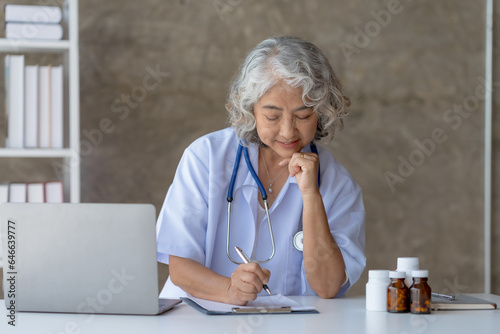 Senior Asian female doctor sitting on desk with laptop and smartphone.