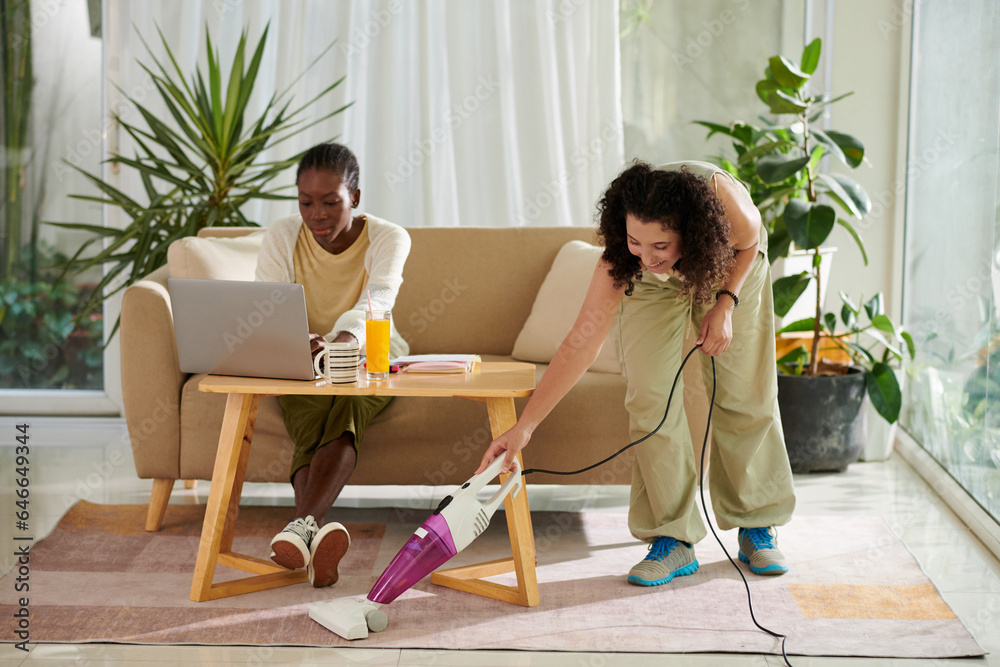 Cheerful girl vacuum cleaning carpet when her roommate studying for exam