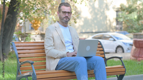 Modern Casual Man Looking at Camera while Working on Laptop Outdoor