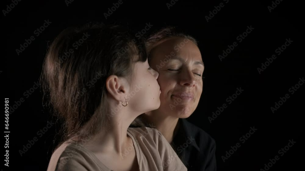 Joyful family sitting against black background. A view of joyful good looking mother hugs her daughter with love against black background. Joyful family sitting against black background. A view of joyful good looking mother hugs her daughter with love against black background.