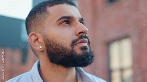 Close up view of serious handsome bearded man with little earring dressed in blue shirt looking at sky and dreaming. Young pensive man of arabian nationality thinking of own purposes in life. photo