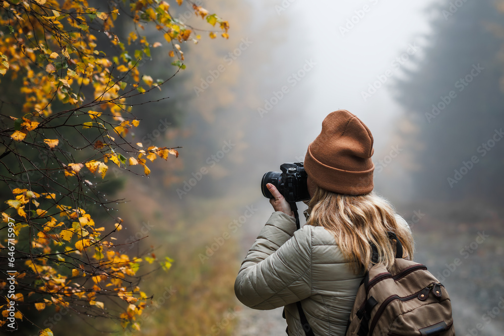 Woman with camera taking picture of autumn leaf. Tourist hiking in misty forest. Landscape photographer with backpack enjoying nature in fall season - obrazy, fototapety, plakaty 