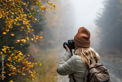 Woman with camera taking picture of autumn leaf. Tourist hiking in misty forest. Landscape photographer with backpack enjoying nature in fall season photo