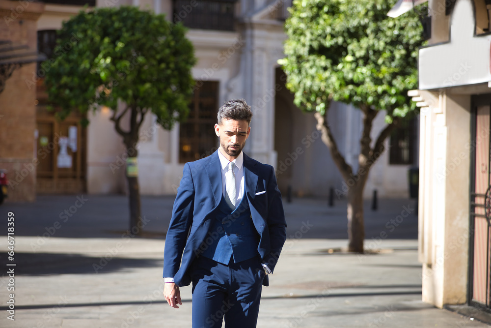 Young and attractive businessman, with beard and suit, walking along a city street with his hand in his pocket. Concept beauty, fashion, success, achiever, trend.