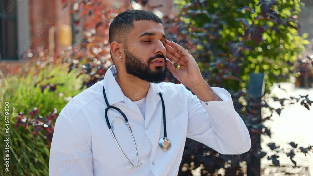 Exhausted male physician getting stress at work and feeling strong headache. Indian young man in medical outfit relaxing on fresh air and massaging temples to get rid of pain. Health concept. Exhausted male physician getting stress at work and feeling strong headache. Indian young man in medical outfit relaxing on fresh air and massaging temples to get rid of pain. Health concept.