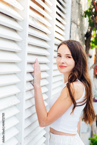 Beautiful young woman interior designer is looking at her project with a happy face. Attractive brunette with long hair in white casual clothes is touching exterior decorative white wall