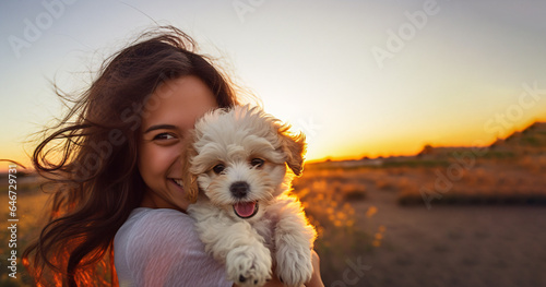 Smiling teen girl holding her cute white maltese puppy outside in the nature, sunset in the background. Loving bond between human and animal. 