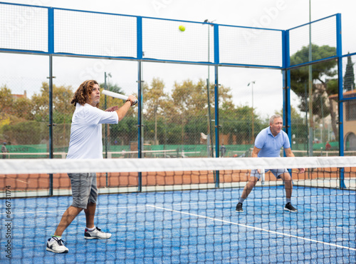 Portrait of adult man enjoying padel racket sport, playing doubles game with elderly male friend on court outdoor © JackF