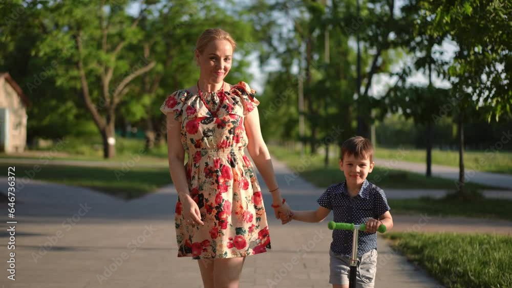Dolly shot portrait of smiling Caucasian woman and handsome little boy looking at camera walking on sunny park alley with scooter. Front view confident mother and son posing enjoying leisure at sunset Dolly shot portrait of smiling Caucasian woman and handsome little boy looking at camera walking on sunny park alley with scooter. Front view confident mother and son posing enjoying leisure at sunset