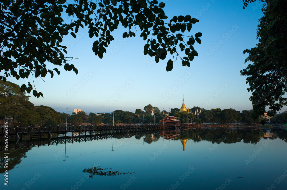 Golden Chan Tha Ya Pagoda and long teak bridge reflecting in calm water of Thinga Yazar Channel, Mandalay, Myanmar