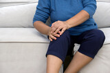 A distressed woman experiencing sharp knee pain seated on a comfy sofa, her hand tenderly resting on the sore spot.
