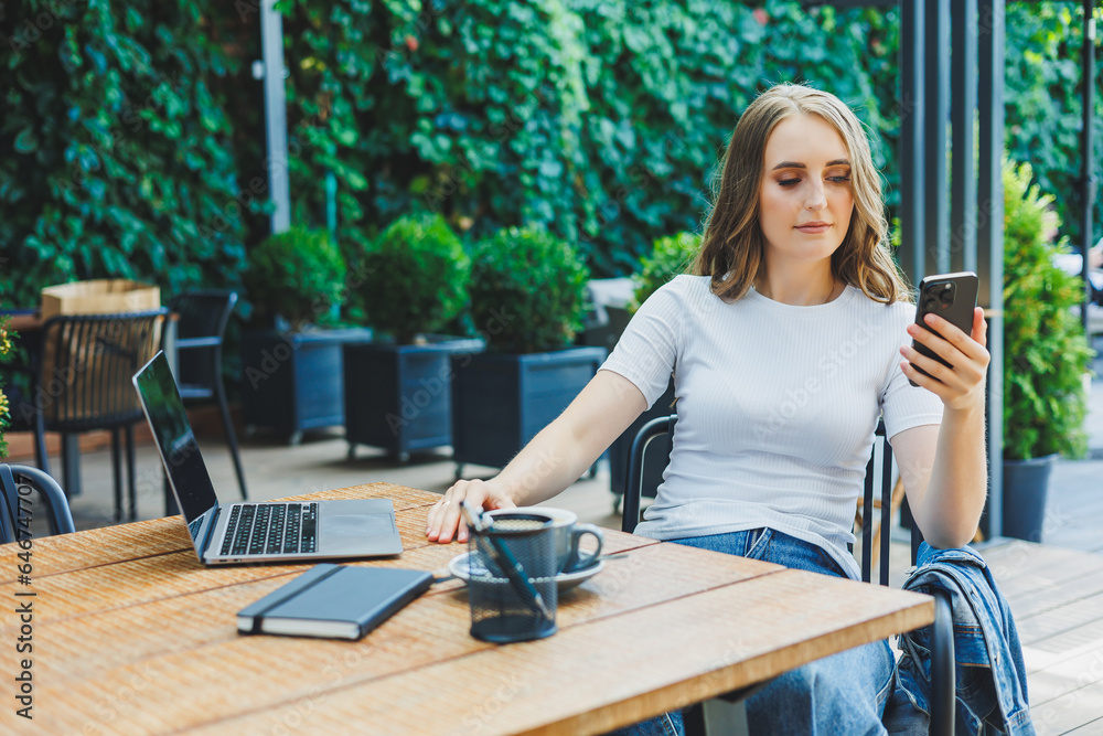 Young woman sitting on the terrace of a cafe on the street of a modern cafe. Woman sitting at table with phone and using laptop outdoors. Remote work in a summer cafe Young woman sitting on the terrace of a cafe on the street of a modern cafe. Woman sitting at table with phone and using laptop outdoors. Remote work in a summer cafe