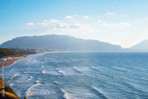 Wonderful view of beach with buildings seashore coastline, sea waves, mountains surrounded by clouds on sunny windy day.