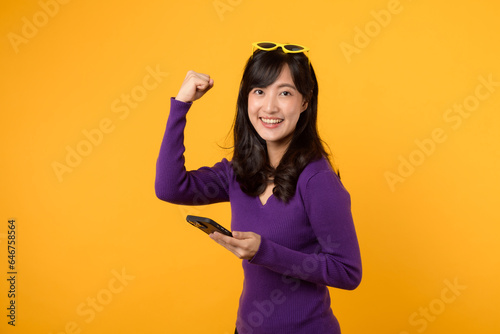 Portrait beautiful young asian woman 30s happy smile dressed in purple sweater with smartphone and fist up hand gesture isolated on yellow studio background. app smartphone concept