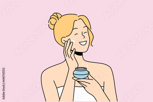 Woman applies anti-aging cream to face to stay beautiful, standing in bath towel after shower. Happy blonde girl smiles and uses cosmetic cream to help prevent wrinkles and pimples.