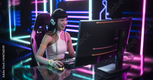 Young Woman Wearing Futuristic White Clothes and Neon Rings, Playing Video Games on a Desktop Computer. Beautiful Girl in Headphones Talking to Online Stream Audience. Streaming and Gaming Concept © Gorodenkoff