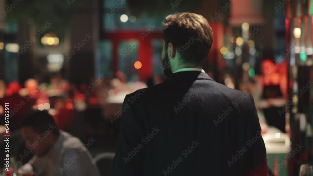 Back view of good-looking gentleman wearing black stylish suit walking confidently among round tables with guests in fancy restaurant. Male wedding host leading show for newlyweds. Back view of good-looking gentleman wearing black stylish suit walking confidently among round tables with guests in fancy restaurant. Male wedding host leading show for newlyweds.
