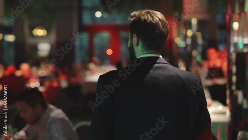 Back view of good-looking gentleman wearing black stylish suit walking confidently among round tables with guests in fancy restaurant. Male wedding host leading show for newlyweds.